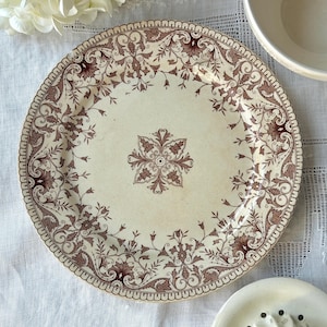 May include: A cream-colored antique plate with a brown floral pattern. The plate has a scalloped edge and a central floral medallion. The design includes leaves and flowers, creating a vintage aesthetic. The plate is part of a tableware set.