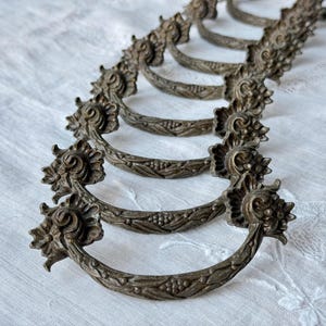 Könnte beinhalten: Ein Satz antiker Schubladengriffe aus Bronze. Jeder Griff hat einen gebogenen Griff mit floralen und Blattdetails. Die Griffe sind auf einer weißen, strukturierten Oberfläche angeordnet, die ihr verziertes Design und ihre gealterte Patina hervorhebt.