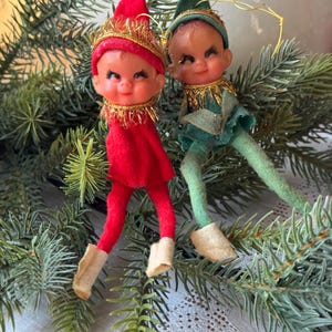 May include: Two vintage Christmas elf ornaments. One elf is dressed in red with a gold crown and the other in green. Both have smiling faces, felt outfits, and beige boots. They are posed on a bed of evergreen branches.