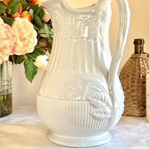 May include: White ceramic pitcher with a fluted design and a leaf motif. The pitcher has a wide mouth and a handle that is shaped like a leaf.