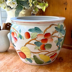 May include: A white ceramic planter with a wide, rounded body and a flared rim. The exterior features a hand-painted design of brown branches with green leaves and various fruits in shades of red, orange, and yellow, including apples and pears.