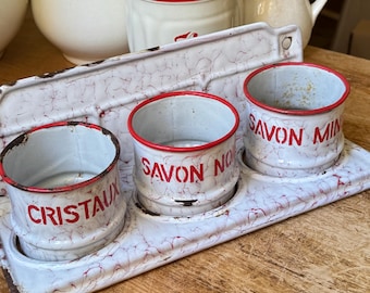 May include: A set of three white enamelware containers with red trim and text, set on a matching tray. The containers are labeled "CRISTAUX", "SAVON NO", and "SAVON MIN". The tray and containers have a distressed, crackled finish, suggesting an antique aesthetic.
