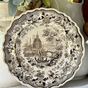 May include: A round ceramic plate with a scalloped edge featuring a dark brown transferware pattern on a cream background. The central design depicts a domed building, a bridge, and a boat with figures, surrounded by a floral border.