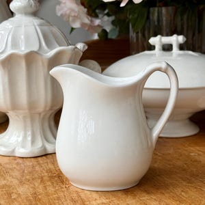 May include: A white ceramic creamer with a curved handle and a pouring spout. The creamer is part of a white ceramic serving set, with other pieces visible in the background. The set is placed on a wooden surface.