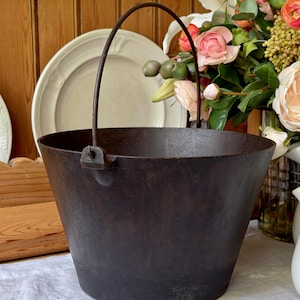 May include: A dark brown, tapered cast iron pot with a smooth, wide opening and a thin, arched metal handle attached to two small side lugs. The exterior surface of the vessel shows a matte, weathered texture consistent with heavy metal cookware.