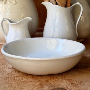 May include: A shallow, round, white ceramic bowl with a smooth, glossy finish and a slightly irregular rim. The bowl is positioned in the foreground, with two white ceramic pitchers and wooden kitchen utensils visible in the background.