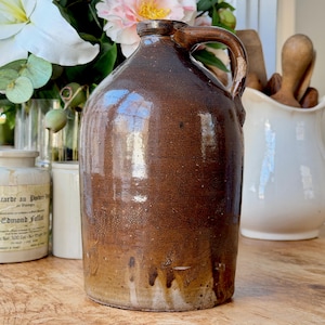May include: A brown stoneware jug with a handle. The jug has a glossy finish with darker brown streaks and drips near the base. The background includes flowers and other antique items.