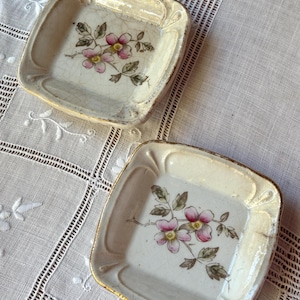 May include: Two square ceramic dishes with a cream-colored glaze and a visible crackle finish. Each dish features a central hand-painted floral motif with pink blossoms, yellow centers, and green leaves, finished with a thin gold-toned rim.
