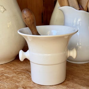 May include: A white ceramic mortar with two small side handles and a wide, flared rim. A dark brown wooden pestle is positioned inside the mortar, resting against the interior wall. The mortar has a smooth, cylindrical base and a glossy finish.