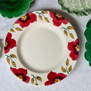 May include: A round ceramic dinner plate with a cream-colored center and a wide rim decorated with a repeating pattern of large red flowers with yellow centers and dark red accents, interspersed with small green and brown leaves.