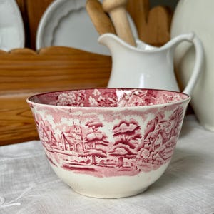 May include: A ceramic bowl featuring a deep red transferware pattern on a cream background. The design depicts a detailed landscape with houses, trees, and a church tower. The interior rim also displays a floral pattern in the same red color.