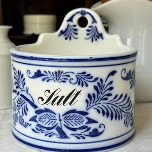 May include: A white ceramic wall-mounted salt cellar featuring a cylindrical body with a flat back and a hanging hole. The exterior is decorated with intricate blue floral and leaf patterns and the word Salt written in a black cursive script.