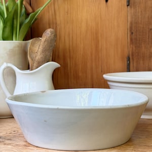 May include: A shallow, off-white ceramic bowl with a smooth, slightly tapered rim and subtle surface variations. The bowl features a clean, minimalist design with a neutral color palette, showcasing a simple, functional shape for kitchen use.