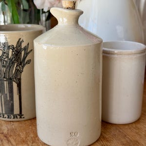 Puede incluir: Colección de cerámica antigua de gres. La pieza central es una botella cilíndrica beige con un tapón de corcho, estampada con el número "20". Otras dos piezas incluyen un tarro cilíndrico y un recipiente más alto con un diseño floral en blanco y negro.