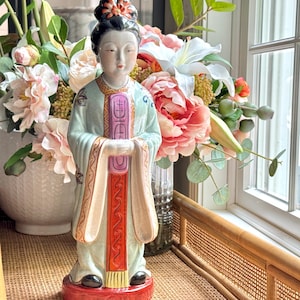 Puede incluir: Figura de cerámica de una mujer con vestimenta asiática tradicional. La figura está pintada en suaves colores pastel, con una base roja y un pergamino morado. La mujer tiene un elaborado peinado y sostiene un pergamino.