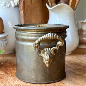 May include: A cylindrical, tarnished brass container with a decorative handle featuring a goat's head. The container has a slightly flared rim and sits on a wooden surface. Other antique kitchenware is visible in the background.