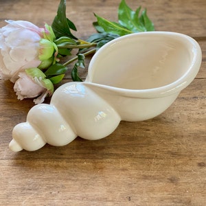 May include: White ceramic seashell planter with a smooth, glossy finish. The planter is shaped like a spiral seashell and has a wide opening at the top.