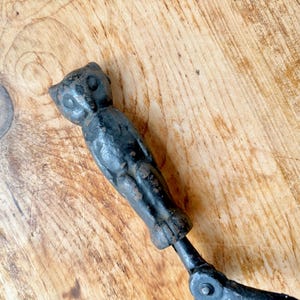 Pode incluir: Uma ferramenta vintage em metal preto em forma de coruja, possivelmente um quebra-nozes ou utensílio de cozinha semelhante. A figura da coruja é detalhada com olhos e penas visíveis. A ferramenta mostra sinais de idade e desgaste, com uma aparência rústica e antiga.