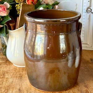 Pode incluir: Um grande pote de grés castanho com um acabamento brilhante. O pote tem duas pequenas alças e texto em relevo perto da borda. Um jarro branco e flores estão no fundo.