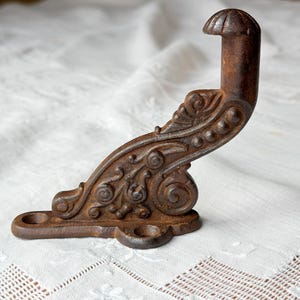 May include: Ornate, antique-style cast iron hook with a dark brown, rust-like finish. The hook features decorative scrollwork and a curved design, perfect for hanging items. The base has two holes for mounting.