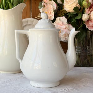 Puede incluir: Una tetera de cerámica blanca con tapa, asa y boquilla. La tetera está sobre un mantel blanco, con una jarra blanca y flores en el fondo. La tetera tiene un cuerpo redondeado y un pequeño pomo en la tapa.