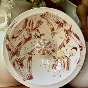May include: A round ceramic lid featuring a cream-colored base with brown botanical illustrations of iris flowers and foliage. The lid has a central knob with a radiating brown and cream striped pattern, providing a decorative finish to the piece.