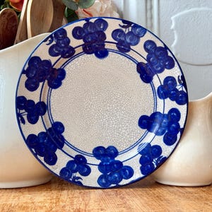 May include: A decorative plate with a white crackle finish and a blue grape design around the rim. The plate is displayed with a white pitcher, a small cream-colored pitcher, and wooden spoons.