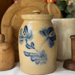 May include: A cylindrical stoneware crock with a speckled tan finish and a matching lid. The exterior features a hand-painted cobalt blue floral design with two prominent blossoms and leafy stems centered on the front of the vessel.