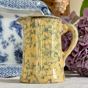 Könnte beinhalten: Ein kleiner, antiker gelber Krug mit Henkel und Ausguss. Der Krug hat ein gesprenkeltes blaues Muster und einen dunkelbraunen Rand. Der Hintergrund umfasst einen blau-weiß gemusterten Teller und lila Blumen.