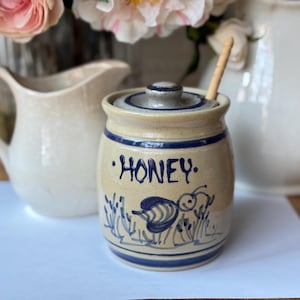 May include: A cream-coloured ceramic honey pot with blue detailing. The word "HONEY" is written in blue script, with a bee and floral design. A wooden honey dipper is inside. The pot has a lid.