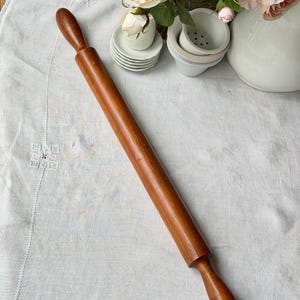 May include: A wooden rolling pin with a dark brown finish. The rolling pin is long and cylindrical, with rounded handles on each end. It rests on a white linen tablecloth, with a floral arrangement in the background.