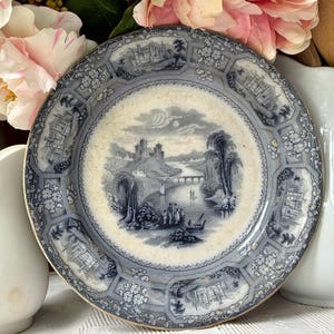 May include: A round ceramic plate featuring a blue and white transferware pattern. The center depicts a river landscape with a castle, bridge, and figures. The border consists of alternating panels showing architectural structures and floral motifs.