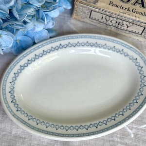 May include: An oval-shaped white ceramic platter featuring a decorative border pattern of small blue diamond shapes. The dish has a smooth, glossy finish and a simple, clean design, providing a neutral base for serving various food items.