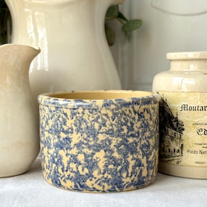May include: A small, round, antique stoneware crock with a speckled blue and cream pattern. The crock has a slightly flared rim and is surrounded by other vintage kitchenware.