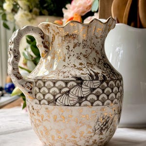 May include: A ceramic pitcher featuring a cream-colored base with gold-leaf splatter accents. The body displays a central band with a black and white fan-shaped pattern and a scalloped rim. It includes a textured handle with gold detailing.