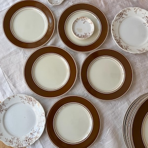 May include: A collection of vintage ceramic plates with a brown border and cream-colored center. Some plates have a floral design. The plates are arranged on a white tablecloth, showcasing a classic tableware set.