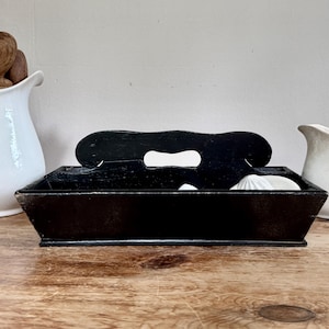 Puede incluir: Una bandeja de madera negra con asa y recorte. La bandeja contiene objetos blancos, posiblemente conchas o elementos decorativos. La bandeja está sobre una superficie de madera, con una jarra blanca y cucharas de madera en el fondo.