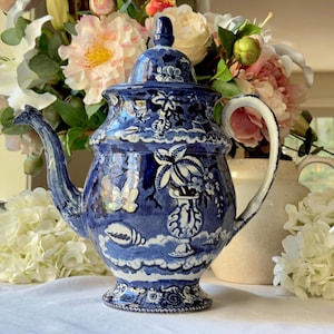 Puede incluir: Una tetera de cerámica azul y blanca con tapa, asa y boquilla. La tetera presenta un detallado diseño floral y de conchas marinas. El fondo incluye flores y un jarrón blanco.