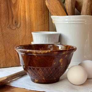 Puede incluir: Un tazón de cerámica esmaltada marrón con un patrón de diamante en la parte inferior. El tazón está sobre una superficie blanca con dos huevos blancos.