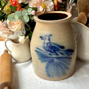 Op de afbeelding: Een beige stenen kruik met een blauwe vogelillustratie. De binnenkant is donkerbruin. De blauwe vogel is afgebeeld op een blauwe tak. De afbeelding bevat bloemen en andere keukenspullen, wat een rustieke uitstraling creëert.