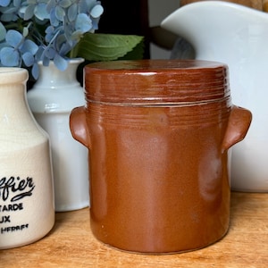 May include: A cylindrical brown glazed ceramic crock with a matching lid and two small side handles. The pot features horizontal grooved lines around the upper body and lid, presenting a smooth, uniform finish throughout the exterior surface.