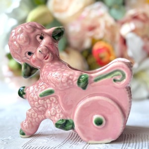 May include: A glossy pink ceramic figurine of a lamb pulling a small cart. The lamb features textured wool, painted black eyes, and green accents on the ears, hooves, and cart wheels. The piece has a hollow opening for small items.