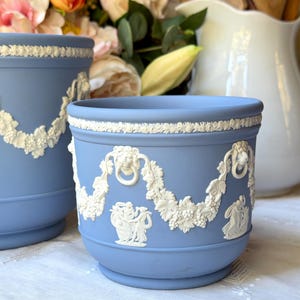 May include: A light blue ceramic planter featuring white relief ornamentation of draped garlands, lion heads with rings, and classical figures. The cylindrical vessel has a smooth matte finish and a raised decorative border along the top rim.