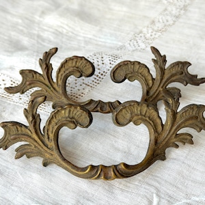 May include: Ornate antique bronze drawer pull with a scrolling leaf design. The hardware features a curved handle and decorative flourishes, perfect for vintage furniture restoration projects.