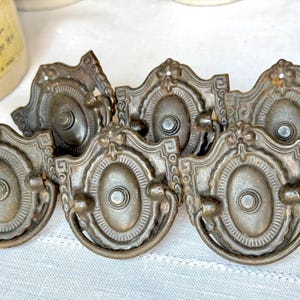 May include: A collection of antique bronze drawer pulls. Each pull features an oval center with a decorative frame and a small handle. The pulls have a vintage, weathered appearance, suggesting age and use. The hardware is likely for furniture restoration.
