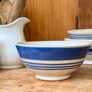 May include: A ceramic bowl with a cream-colored base and a wide blue band around the upper exterior, accented by three thinner blue horizontal stripes below it. The bowl features a rounded rim and a small circular footed base for stability.