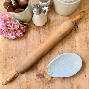 May include: A wooden rolling pin with wooden handles, alongside a white ceramic leaf-shaped dish. The image also includes a pink flower, a small ceramic pitcher, and other kitchenware on a wooden surface.