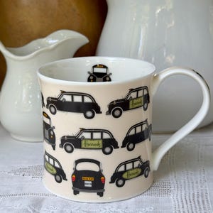 May include: A cream-colored ceramic mug featuring a repeating pattern of black London taxi cabs. Several of the taxis display a green Harrods logo on the side. One taxi is shown from the rear, displaying the license plate F316 PJP and a small 30676.