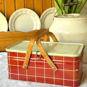 Cesta de picnic de hojalata a cuadros rojos y beige antiguos con asas dobles de madera y cristal.