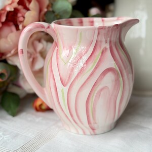 Puede incluir: Una jarra de cerámica rosa y blanca con asa y boquilla. La jarra presenta un patrón de remolino de rayas rosas y verdes. La jarra se utiliza probablemente para servir bebidas.